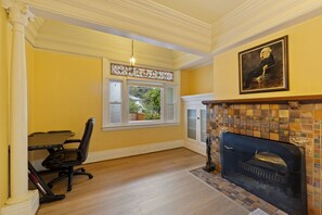 A room with a fireplace, a dining table with chairs, and a window with a view of greenery. - Victorian 3BR with Jetted Tub + Fireplaces - Central District (Seattle)
