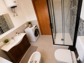 A modern bathroom with a built-in washing machine, a glass shower enclosure, and a separate toilet area. - Between Taormina and the Sea - Casa Rosa (Calatabiano)