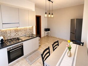 A modern kitchen with a gas stove, oven, and stainless steel appliances. There is a dining area with a table and chairs, and a small seating area with a chair and a table. - Between Taormina and the Sea - Casa Rosa (Calatabiano)