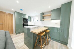A modern kitchen with a central island, wooden stools, and green cabinets. - A Little Home of Your Own in Poulton — Gated Entrance & Parking (Poulton-le-Fylde)