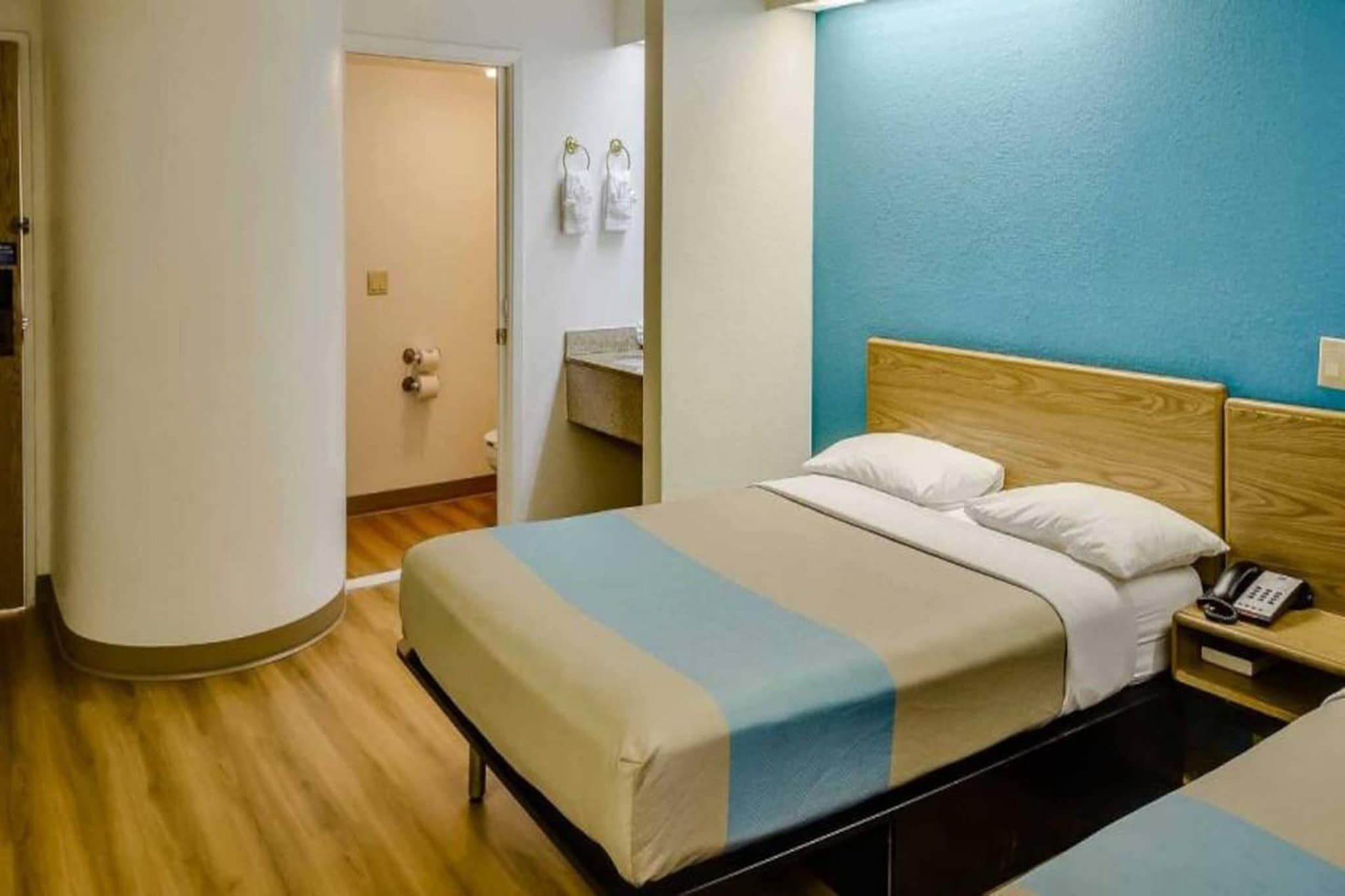 Una habitación de hotel con una cama, una mesita de noche con teléfono y un baño visible a través de una puerta abierta.