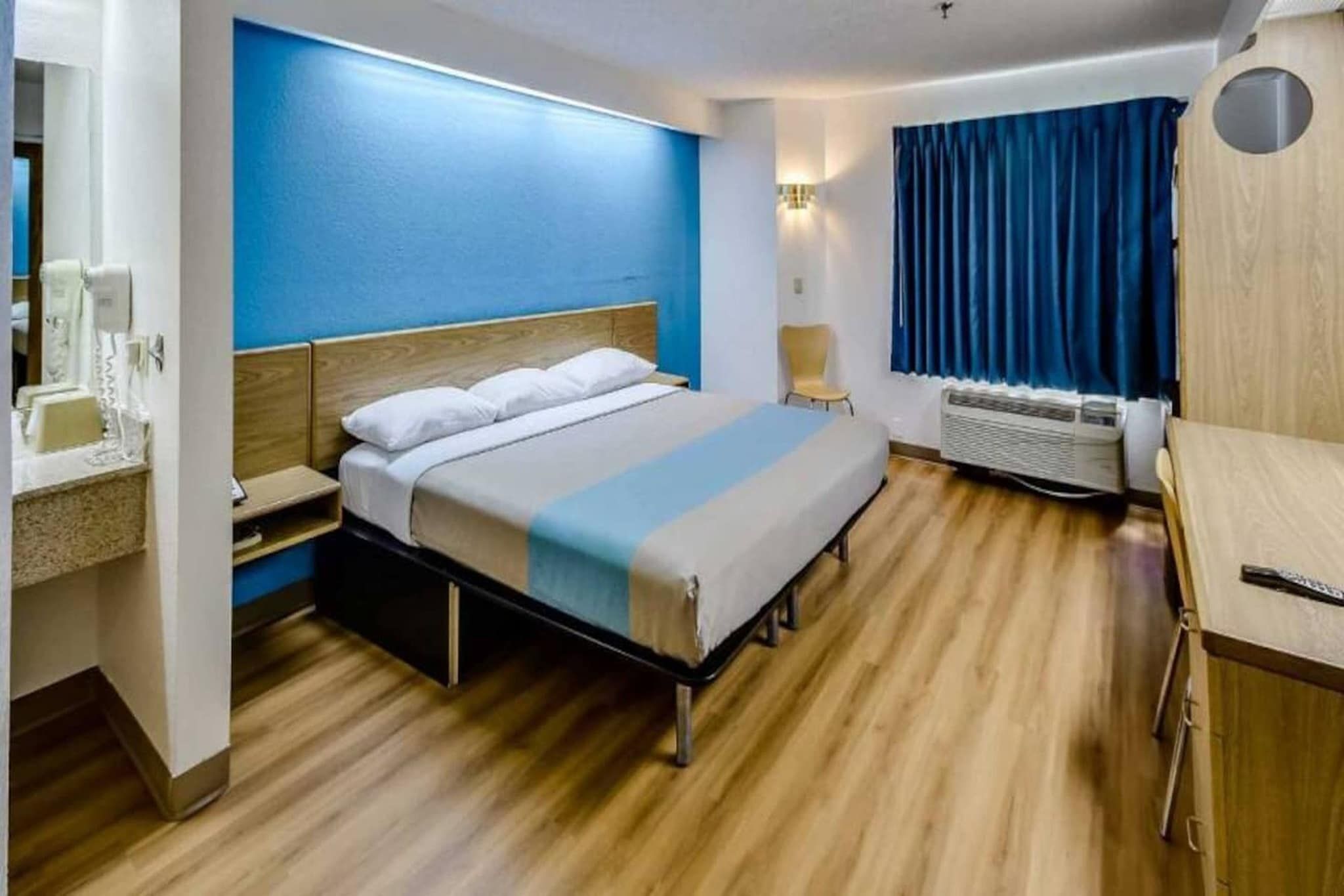 Une chambre d’hôtel comprenant un lit, une table de chevet, un bureau, une chaise, un radiateur et une fenêtre avec des rideaux.