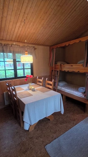 Interior d'una cabana amb una taula de dinar, cadires, llits superposats i una finestra amb cortines.