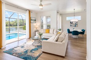 A spacious living room with a large glass door leading to a pool, a white sofa, a glass coffee table, and a dining area with teal chairs. - Lakefront near Disney: Pool + LEGO game room (Kissimmee)