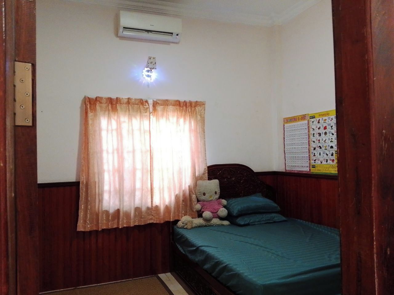 Un dormitorio con cama, un oso de peluche, una ventana con cortinas, un aire acondicionado y un calendario en la pared.