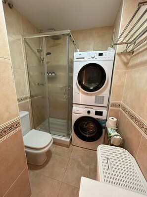 Ein kleines Badezimmer mit einer Duschkabine aus Glas, einer Toilette, einer übereinander angeordneten Waschmaschine und einem Trockner sowie einer kleinen Ablage für Pflegeprodukte.