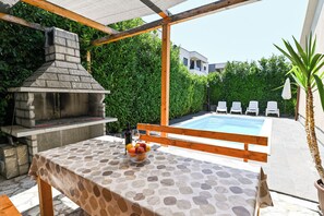 An outdoor patio with a stone barbecue, a table with a patterned tablecloth, a bowl of fruit, and a pool in the background. - Holiday home Lucia 5 minutes’ walk from Old Town in Trogir with private pool (Trogir)
