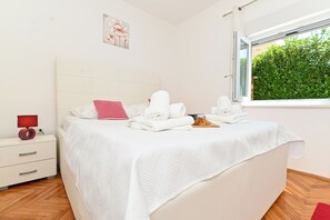 A neatly made bed with white linens and a red pillow, a bedside table with a lamp, and a window with a view of greenery. - Holiday home Lucia 5 minutes’ walk from Old Town in Trogir with private pool (Trogir)