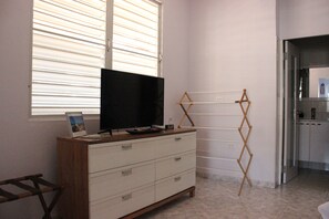 Une pièce avec une commode en bois, un téléviseur posé dessus, un étendoir et un coin cuisine visible à travers une porte ouverte.