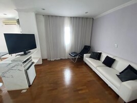 Uma sala de estar com televisão de tela plana, um sofá branco, uma cadeira preta e piso de madeira.
