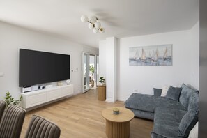A modern living room with a flat-screen TV, a grey sofa, a round wooden coffee table, and a painting of sailboats on the wall. - Casa Zucchero (Cukrići)