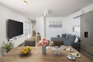 Een moderne woonkamer met een flatscreen televisie, een eettafel met fruit en bloemen, een grijze bank en een schilderij aan de muur.