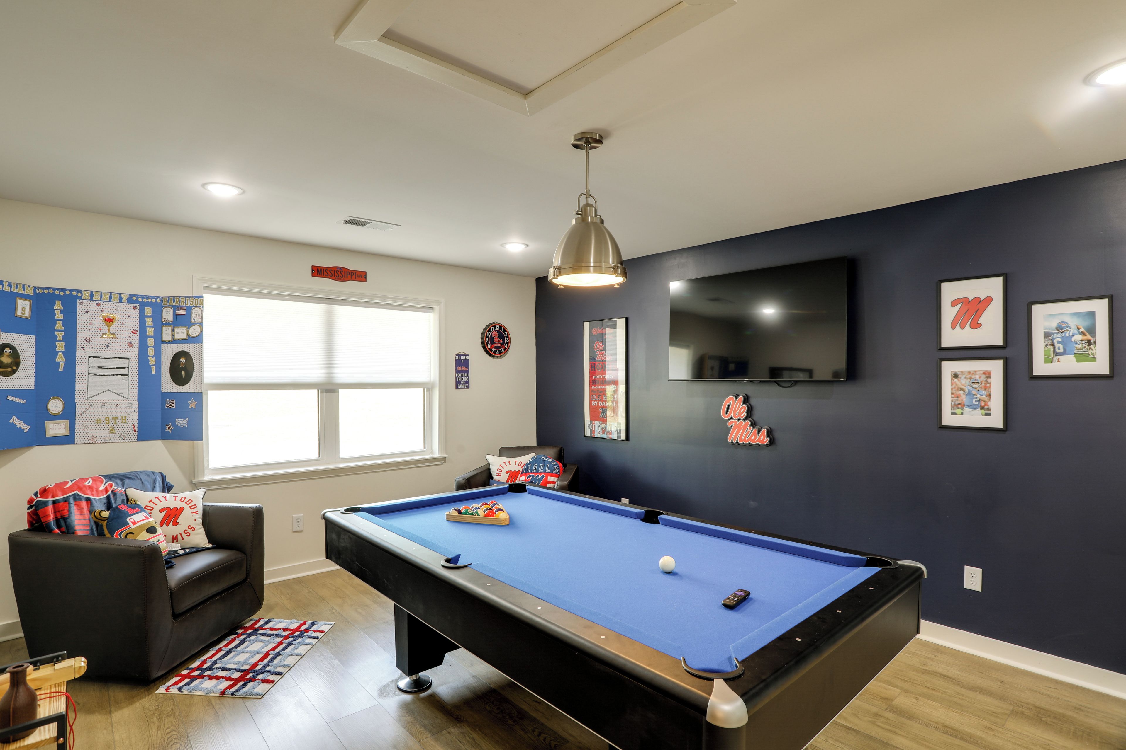 Una habitación con mesa de billar, televisor montado en la pared y una ventana con persianas.