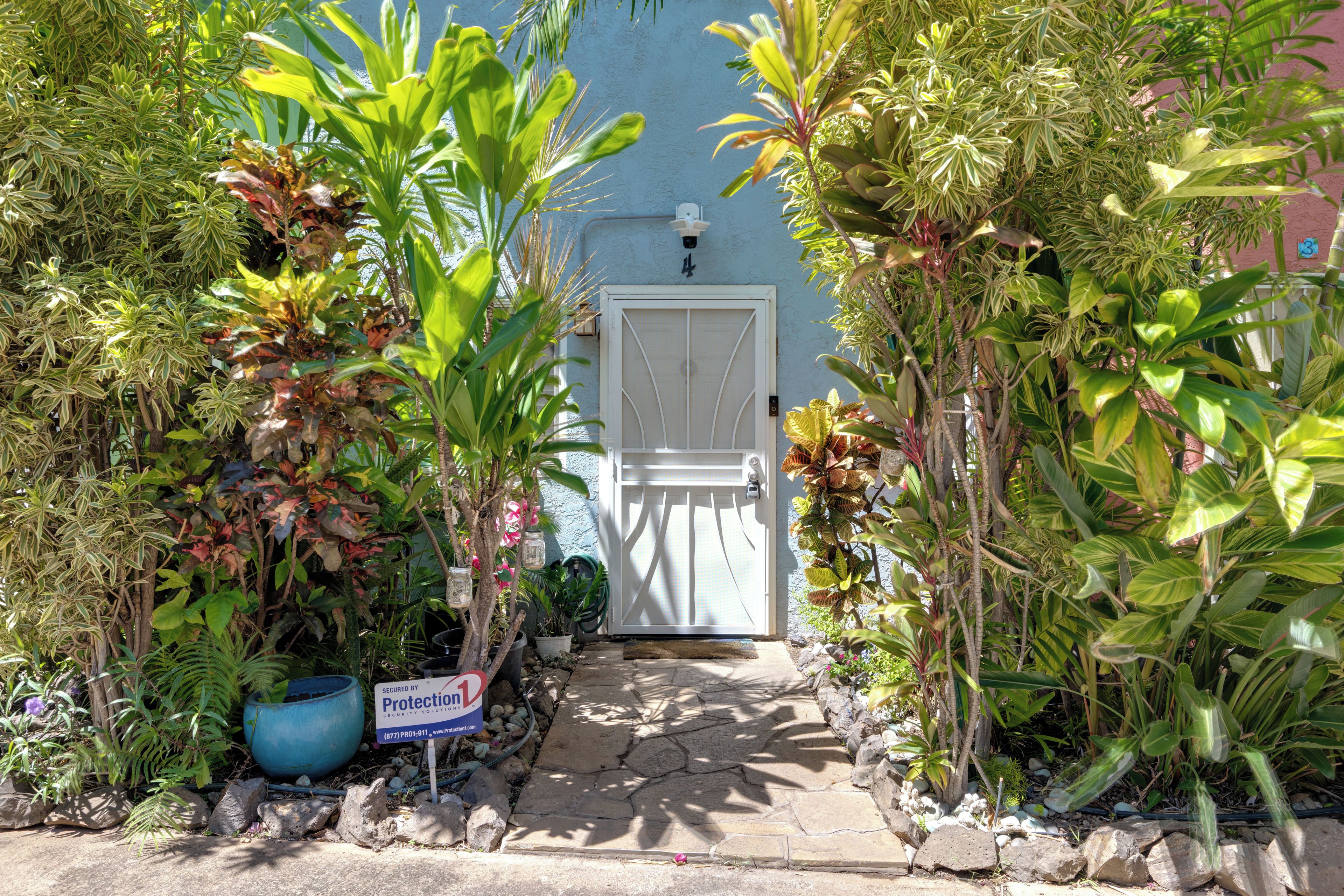 Un cancello con un vialetto in pietra che conduce a una porta bianca, circondata da rigogliosa vegetazione tropicale.