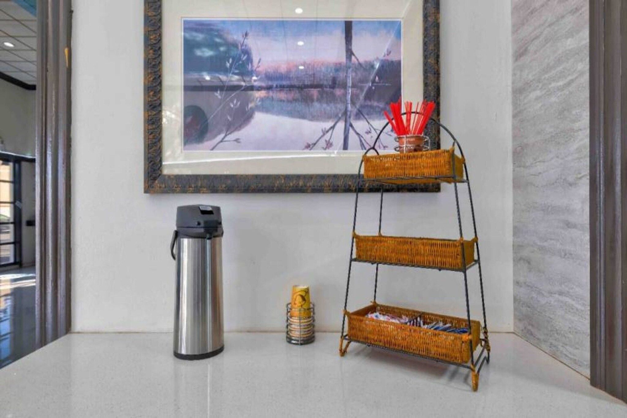 A framed artwork on the wall, a stainless steel thermos, a stack of cans, and a three-tiered wicker shelf with red straws.
