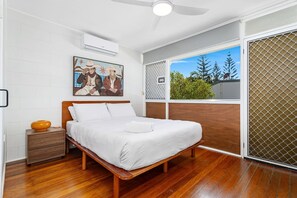 Ein Schlafzimmer mit einem hölzernen Bett, weißer Bettwäsche, einem Nachttisch mit einer Schale, einem Deckenventilator, einem Fenster mit Blick auf Bäume und einer Wand-Klimaanlage.