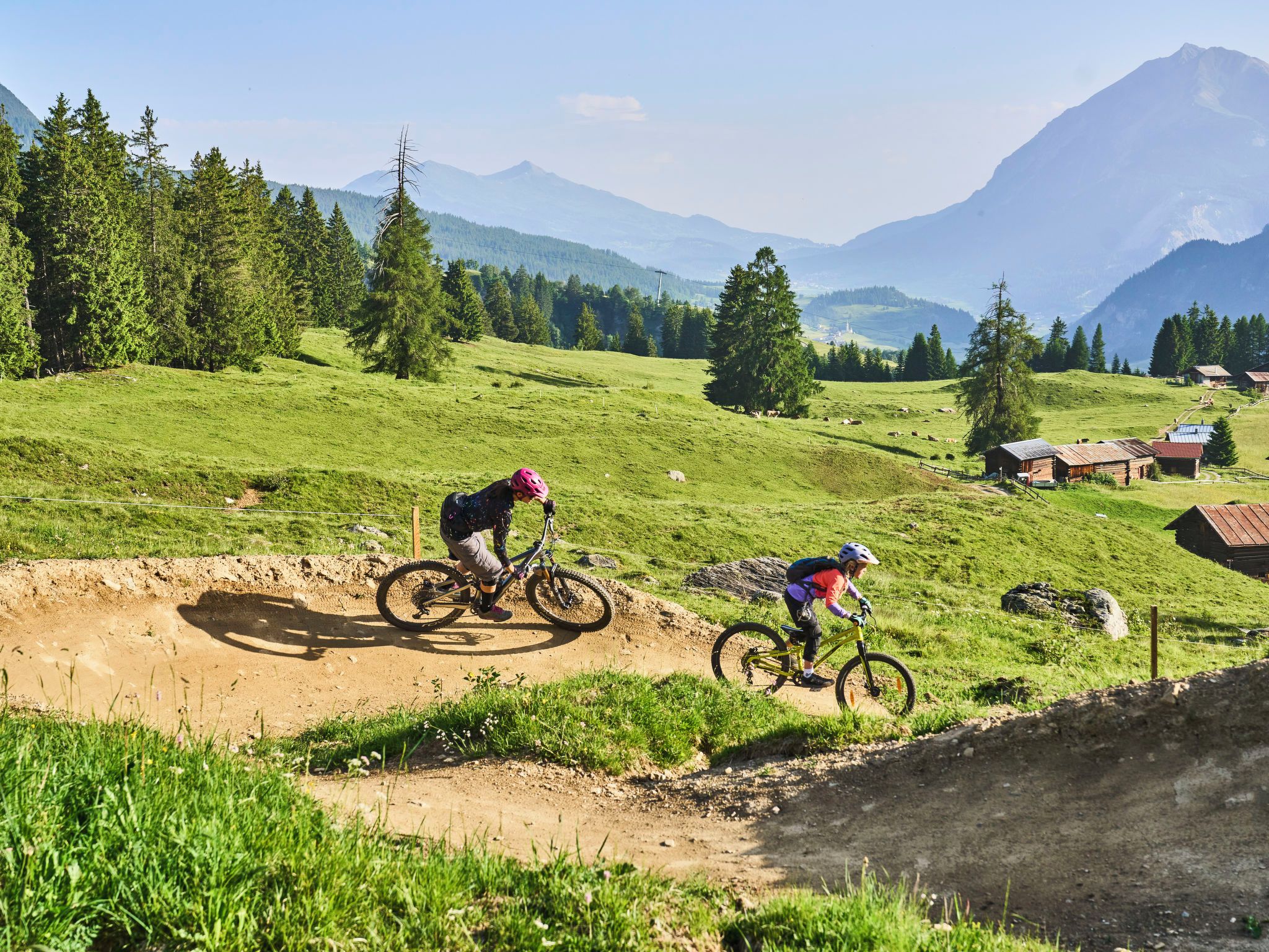 A mountain biking trail with riders, surrounded by green fields and distant mountains.