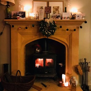 A fireplace with a lit fire, a mantel decorated with cards and a wreath, and a basket on the floor. - Castle in the Cotswolds (Shipston On Stour)