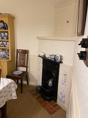A corner room with a fireplace, a wooden chair, a table with a tablecloth, and a shelf with blue and white dishes.