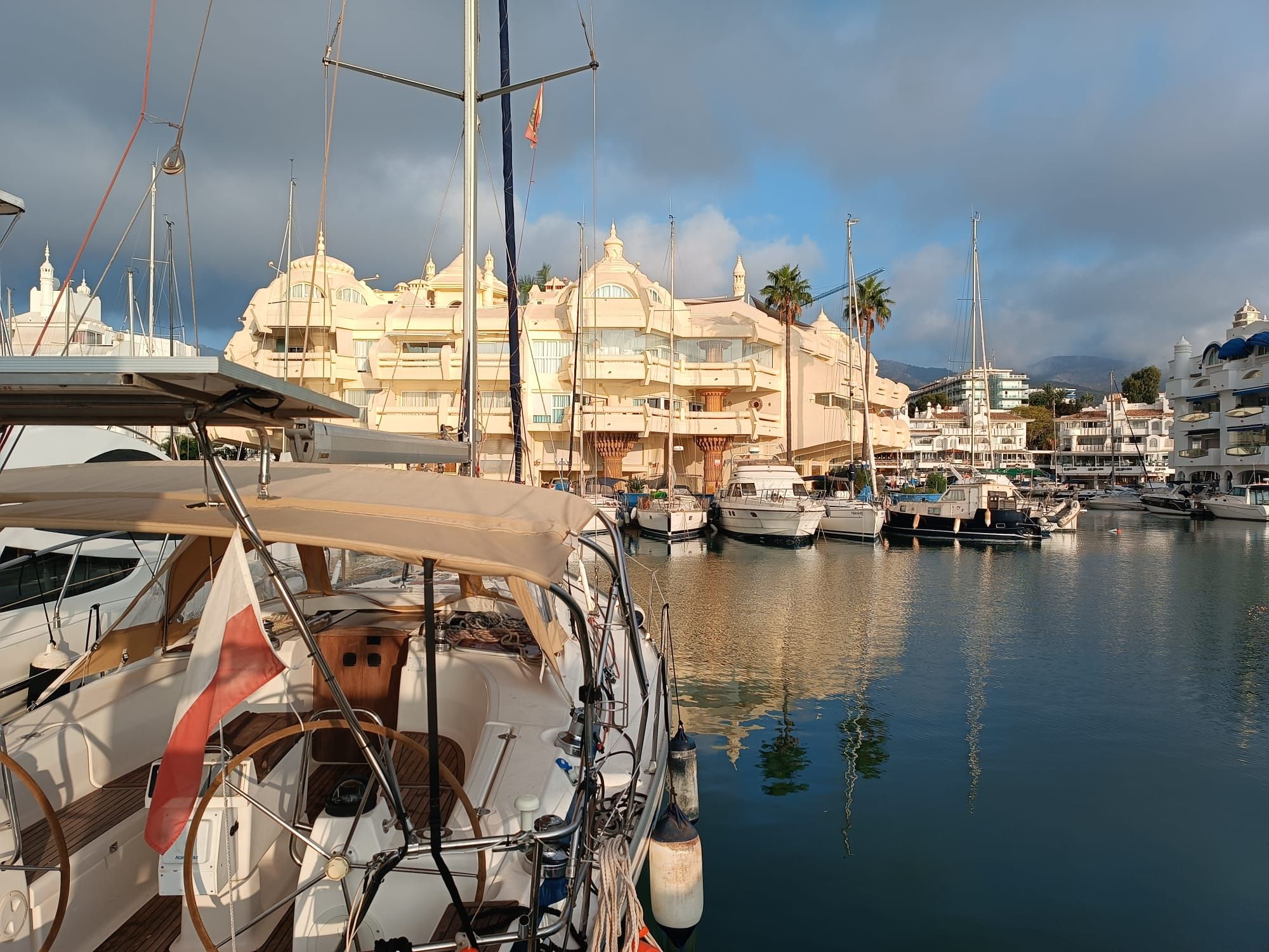 A marina with multiple yachts docked, a large building with a unique architectural design, and calm water reflecting the boats.