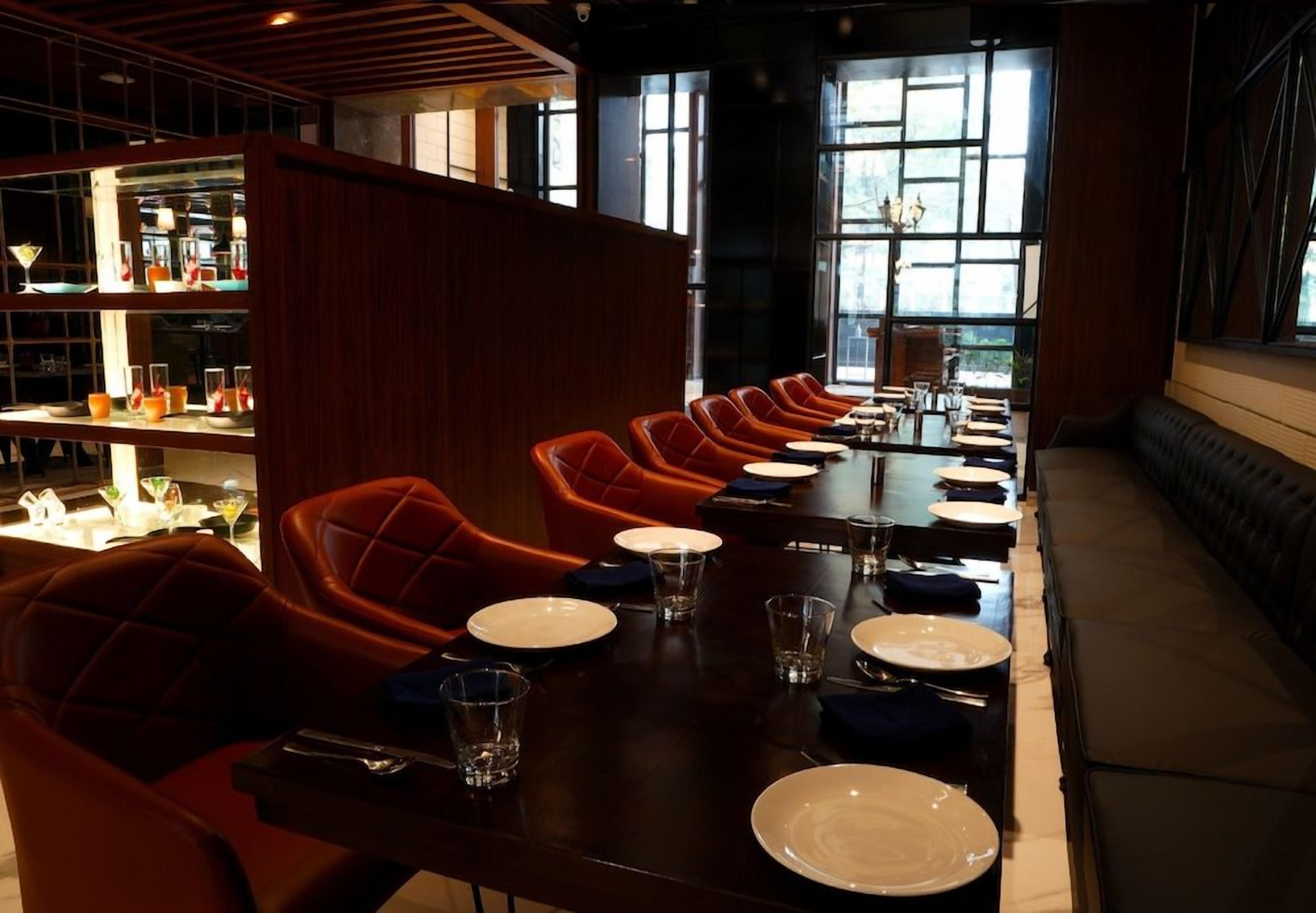 A restaurant with a long table set for dining, red upholstered chairs, and a bar area with shelves of bottles.
