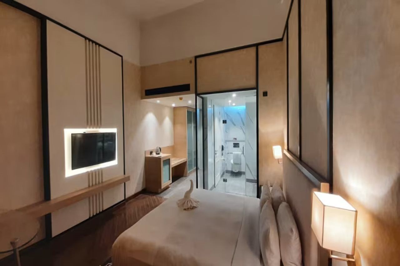 Et hotelværelse med seng, sengebord, lampe og udsigt til et badevrelse gennem en glasdør.