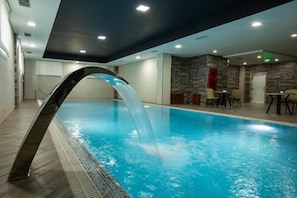 An indoor swimming pool with a water feature, a seating area with tables and chairs, and a stone wall. - Volga Hotel Baku (Baku)