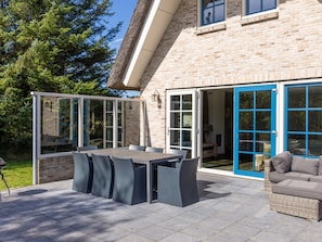 An outdoor patio with a dining table and chairs, a wicker sofa, and a blue-framed sliding door leading to an interior space. - Charming Villa in the Dunes of Ameland for 8 (Nes)