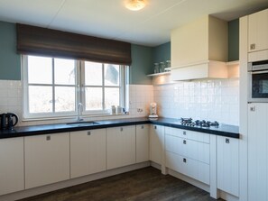 A kitchen with white cabinets, a black countertop, a window with a brown blind, and a built-in oven. - Charming Villa in the Dunes of Ameland for 8 (Nes)