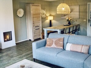 A living room with a sofa, a fireplace, a dining table, and a hanging light fixture. - Charming Villa in the Dunes of Ameland for 8 (Nes)