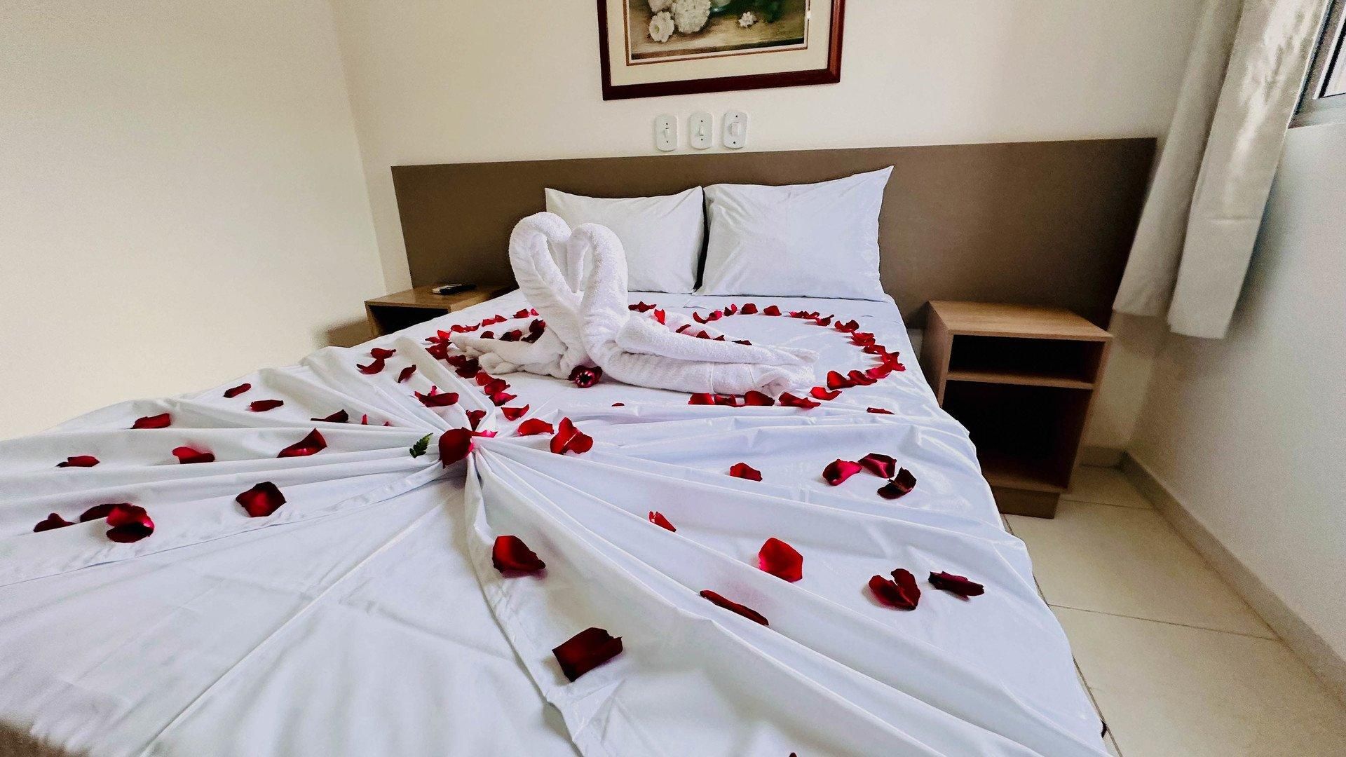 A neatly made bed with rose petals and a towel arranged in a heart shape, a bedside table, a nightstand, and a wall-mounted light switch.