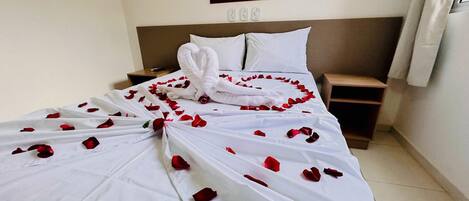 A neatly made bed with rose petals and a towel arranged in a heart shape, a bedside table, a nightstand, and a wall-mounted light switch.