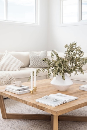 Ein Wohnzimmer mit Couch, Kissen, einem Holztisch, Büchern und einer Pflanze in einer Vase.