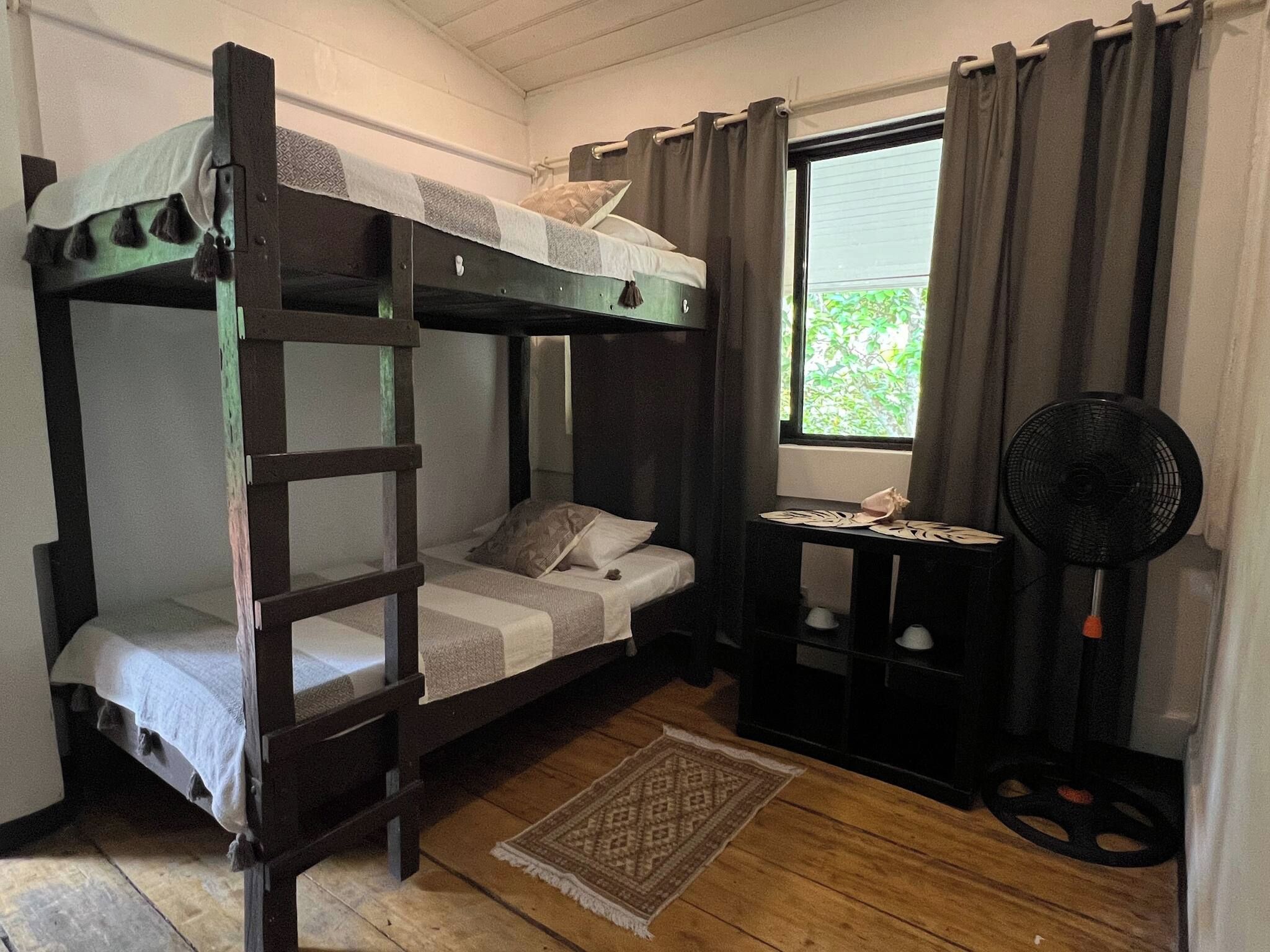 Une chambre avec un lit superposé, une petite table et un ventilateur, une fenêtre avec des rideaux et un tapis sur le sol.