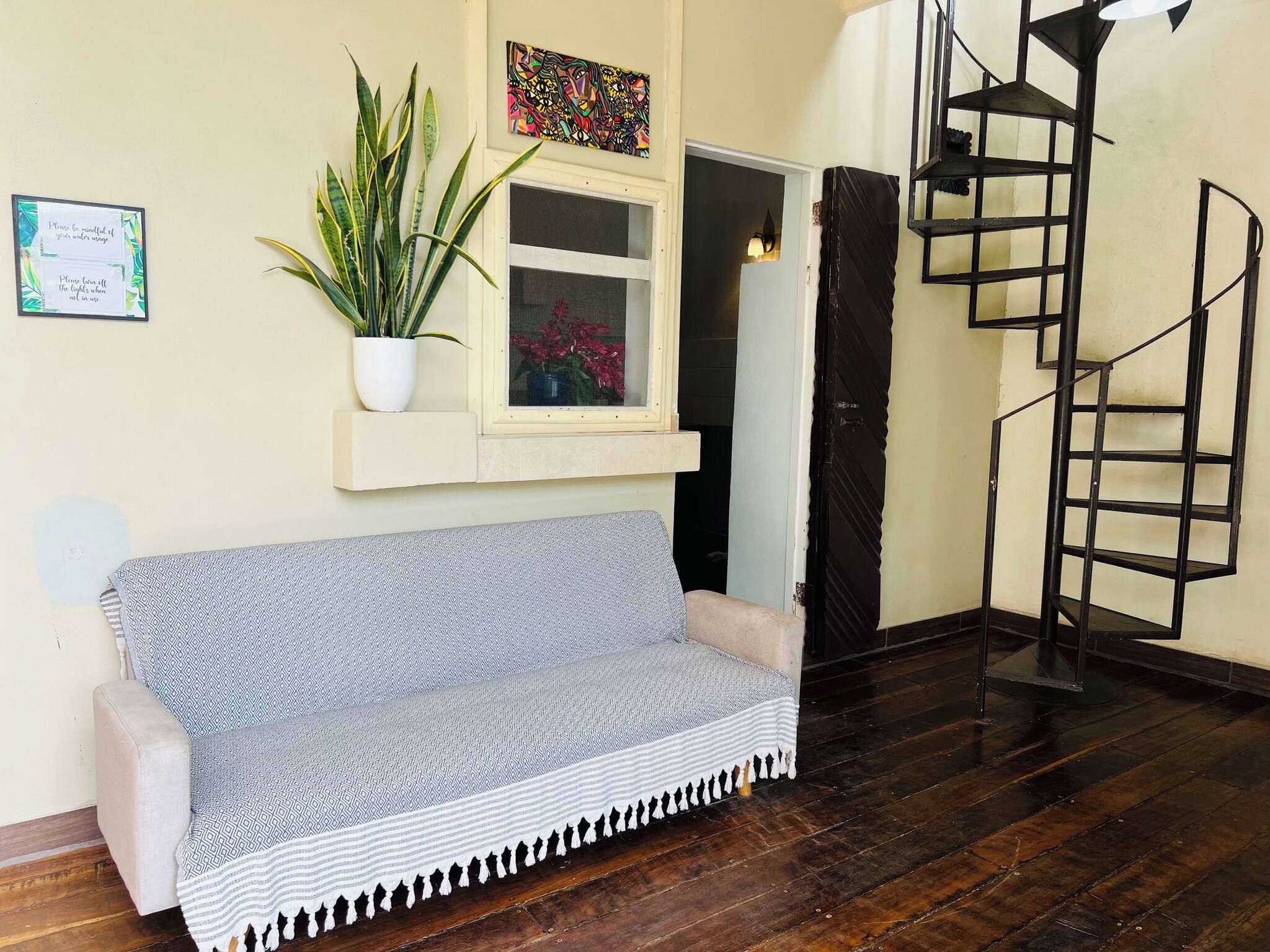 Une pièce avec un canapé, une plante en pot, une fenêtre décor ée de fleurs, un tableau et un escalier en colimaçon.