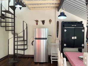 Una cocina compacta con un refrigerador de acero inoxidable, un gabinete negro con detalles decorativos, una escalera de madera y un radiador.