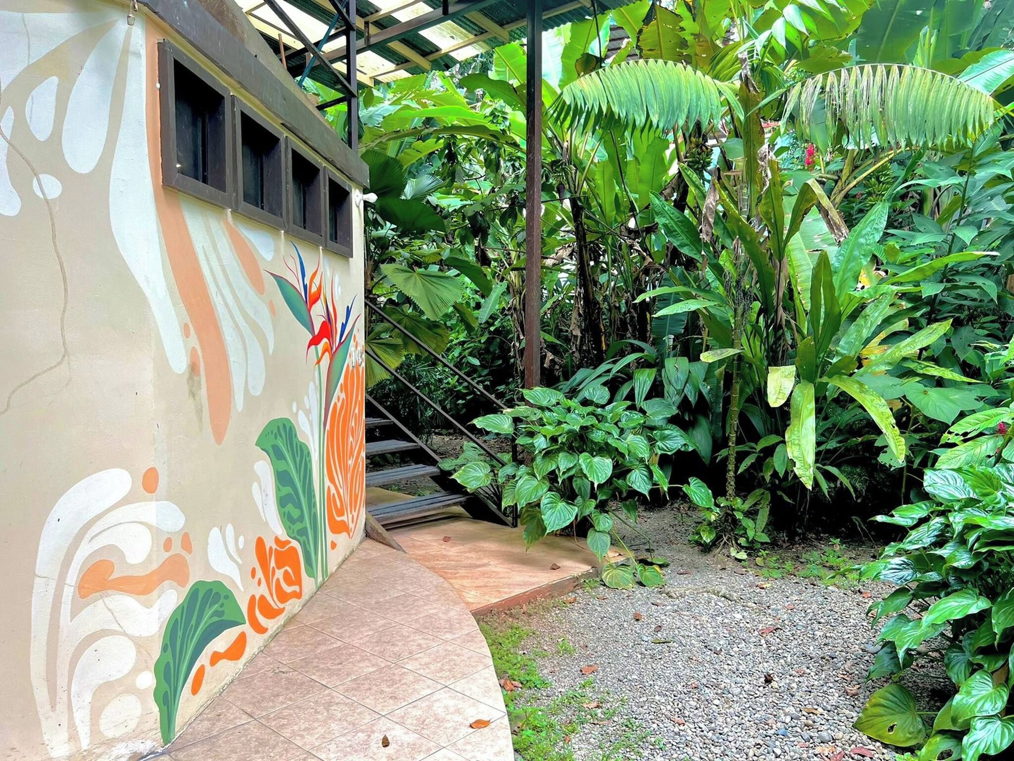 Un espace extérieur abrité, doté d’une allée dallée, entouré d’une végétation luxuriante et bordé d’un bâtiment orné d’une fresque colorée.