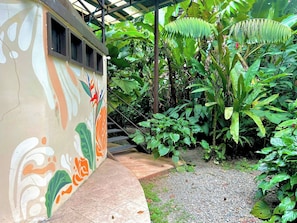 Un espace extérieur abrité, doté d’une allée dallée menant à un escalier, entouré d’une végétation luxuriante et bordé d’un bâtiment orné d’une fresque colorée.