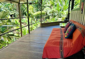 Une terrasse en bois avec un banc et des coussins aux couleurs vives, entourée d’une végétation tropicale luxuriante.