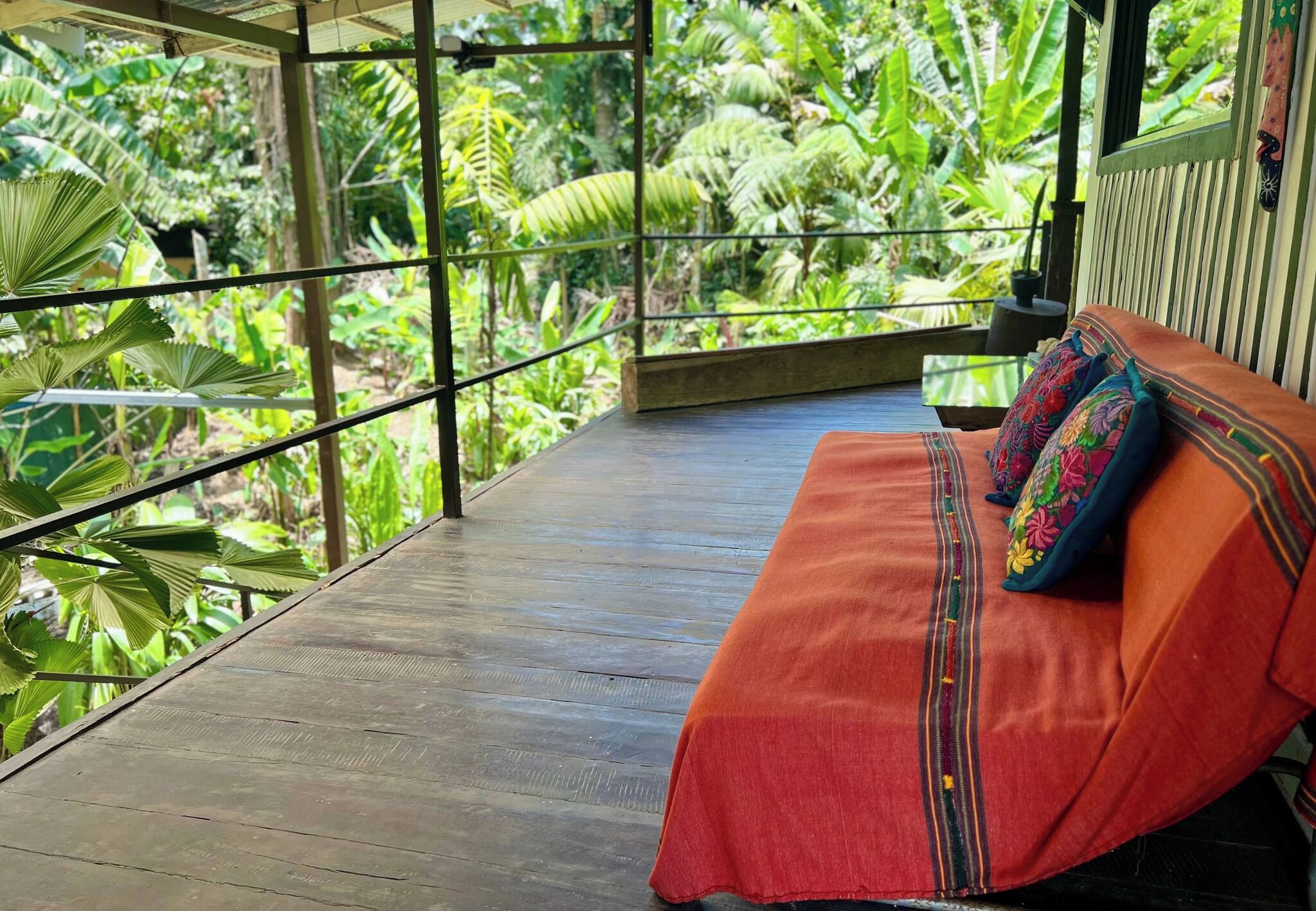 Une terrasse en bois avec un banc et des coussins aux couleurs vives, entourée d’une végétation tropicale luxuriante.