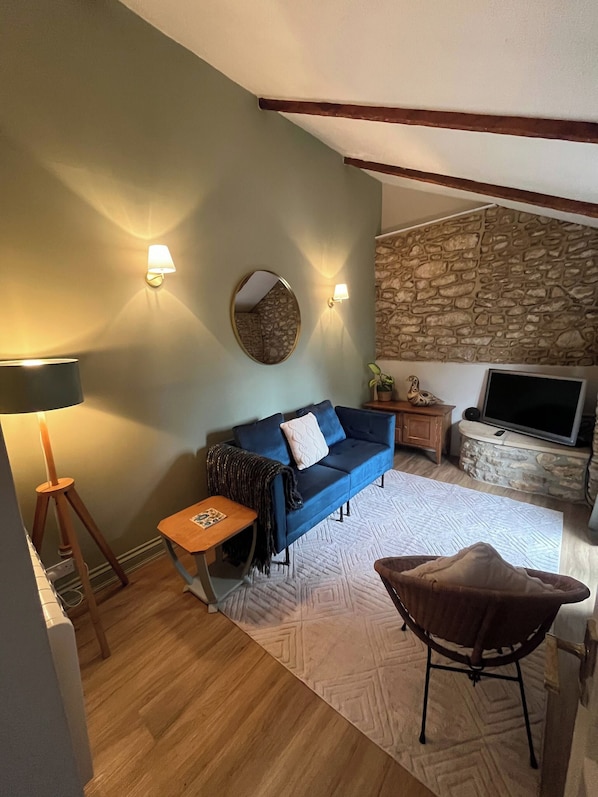 A cozy living room with a blue sofa, a small side table, a lamp, a stone wall, and a television. - Cozy Holiday Retreat in Bradford Abbas - Modern Comfort Awaits (Sherborne)