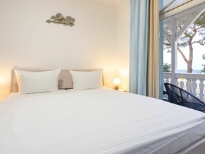 A bed with white linens, a nightstand with a lamp, and a view of the outdoors through a window with a balcony. - No. 7 in Villa Strandburg (Binz)