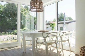 A dining area with a white table and chairs, a woven pendant light, and a view of greenery outside. - Timmare IV Fritz Cabin (Timmendorfer Strand)