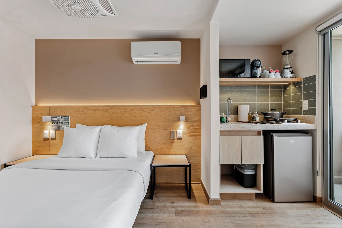 Habitación de hotel con cama, mesita de noche y zona de kitchenette que incluye un refrigerador, cafetera y microondas.