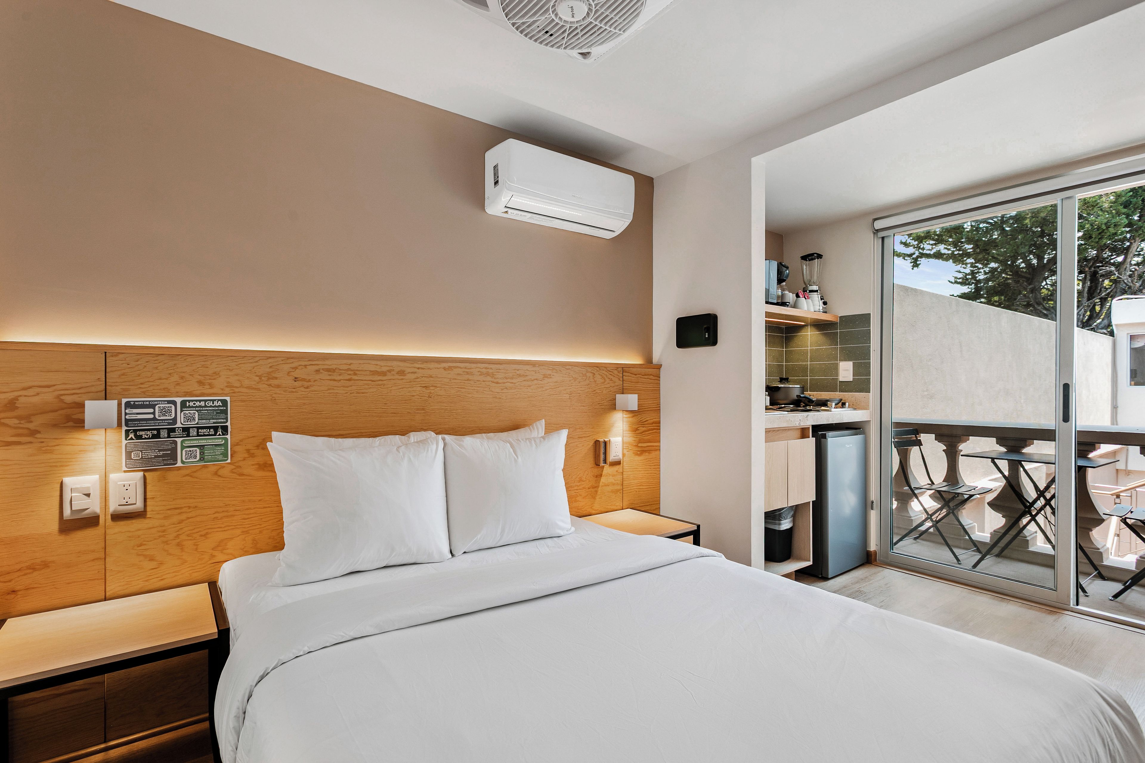 Habitación de hotel moderna con cabecera de madera, cama grande, aire acondicionado integrado, kitchenette y balcón con mobiliario exterior.