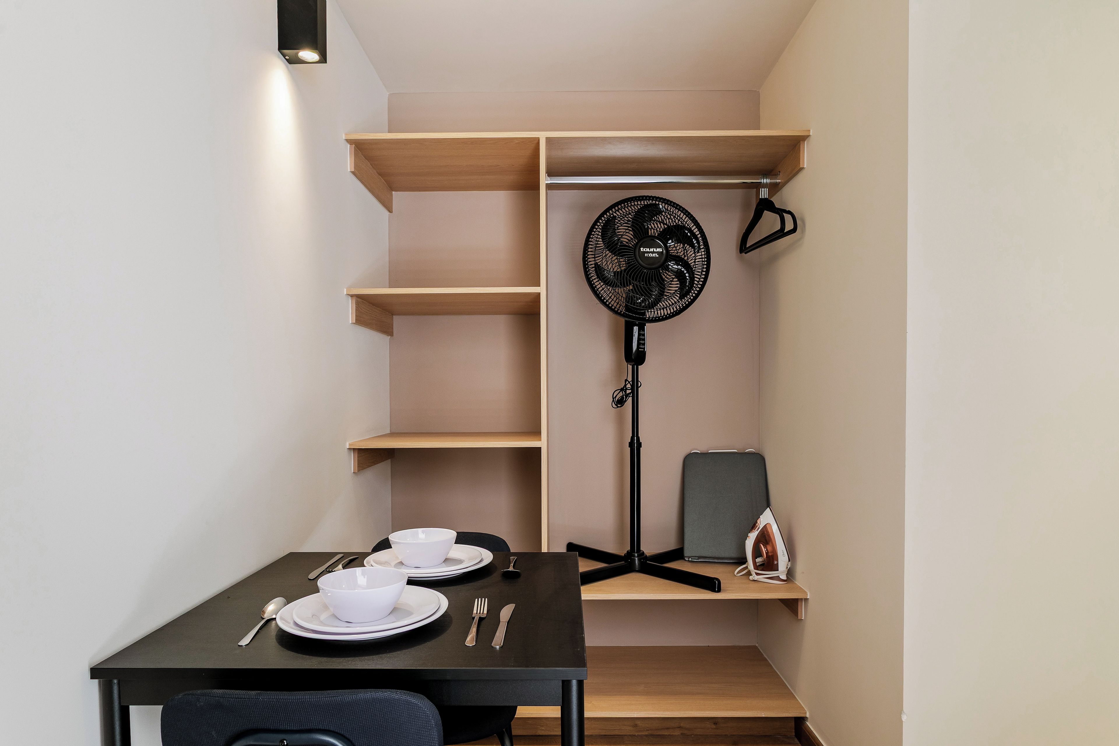Habitación pequeña con una mesa de comedor lista para dos personas, un ventilador de pie y una plancha pequeña.