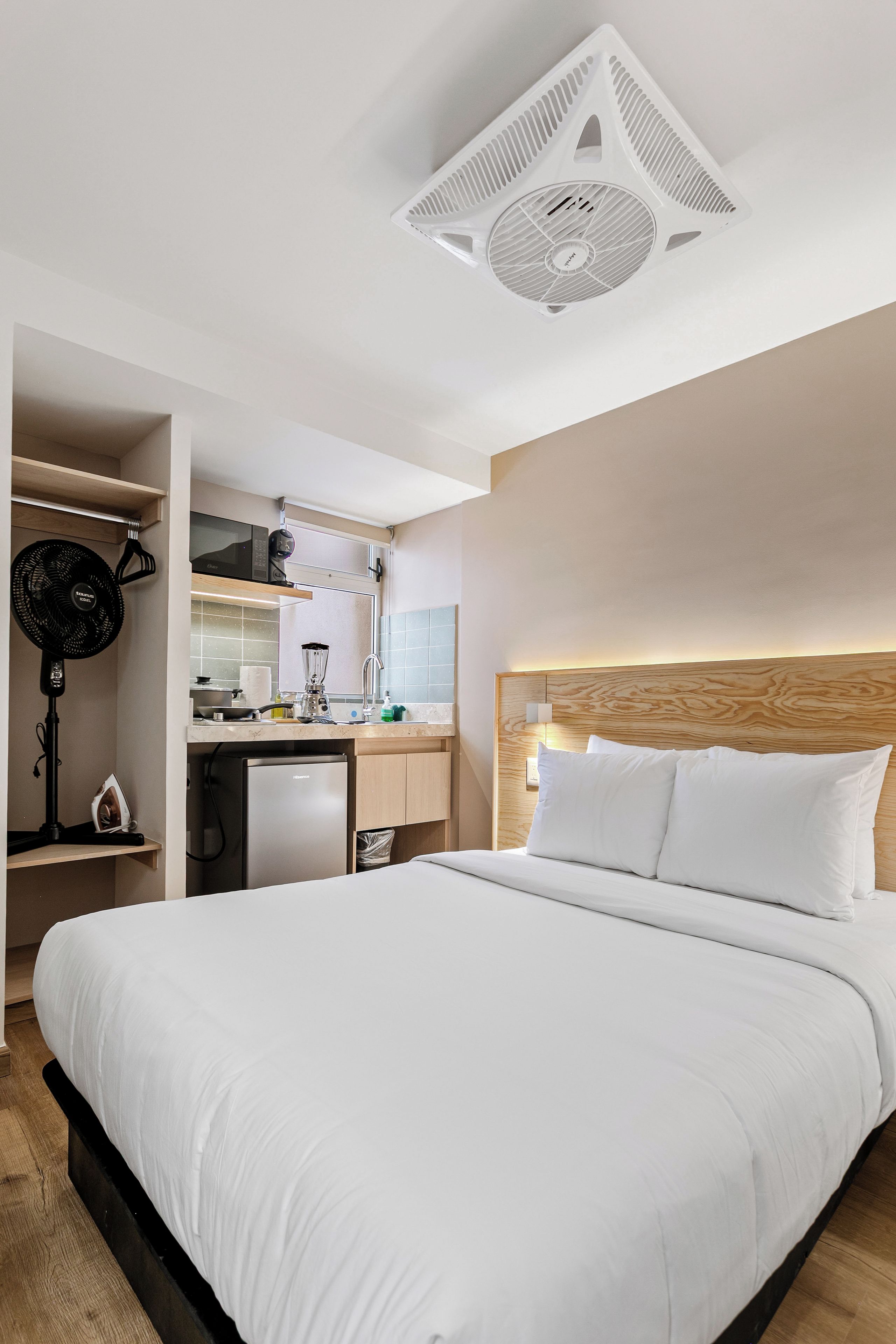 A hotel room with a large bed, a kitchenette, a ceiling fan, and a wooden headboard.