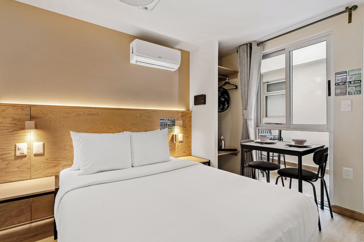 Habitación de hotel con cama, mesita de noche, aire acondicionado de pared y un pequeño escritorio con sillas.