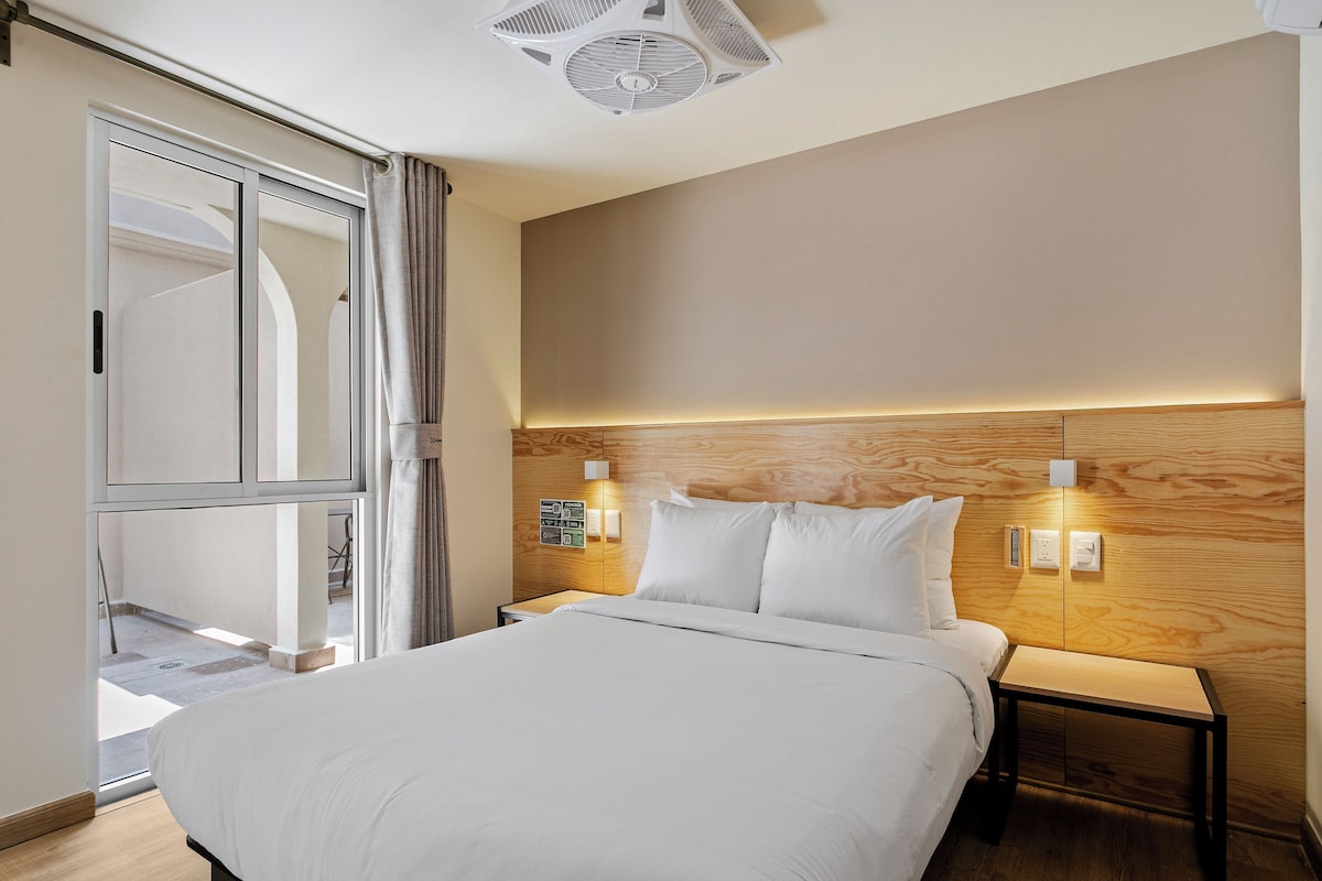 Habitación de hotel con cama, mesita de noche, lámpara de pared y ventilador de techo.