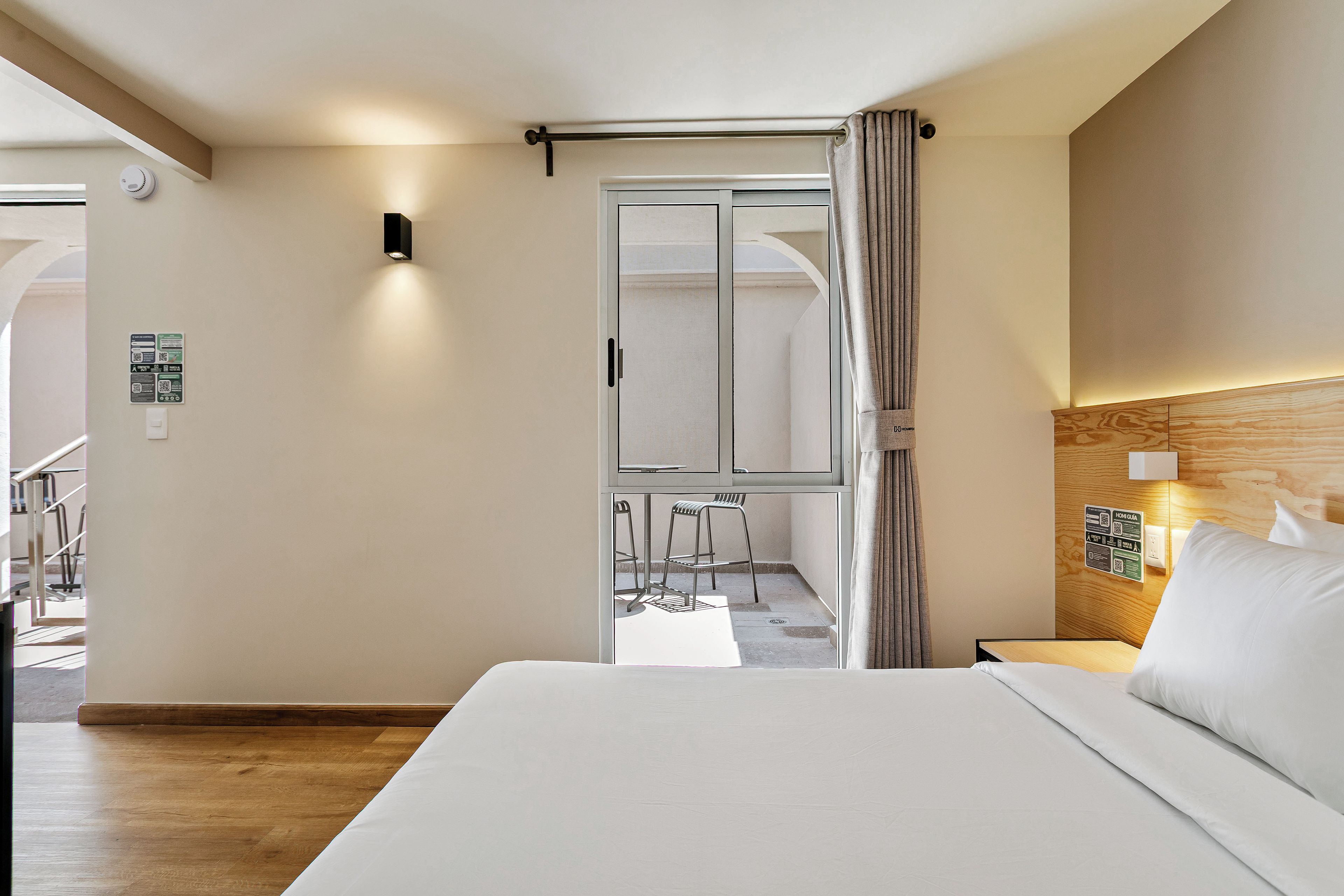 A hotel room with a bed, a nightstand, a wall-mounted light, a window with curtains, and a view of the outside.